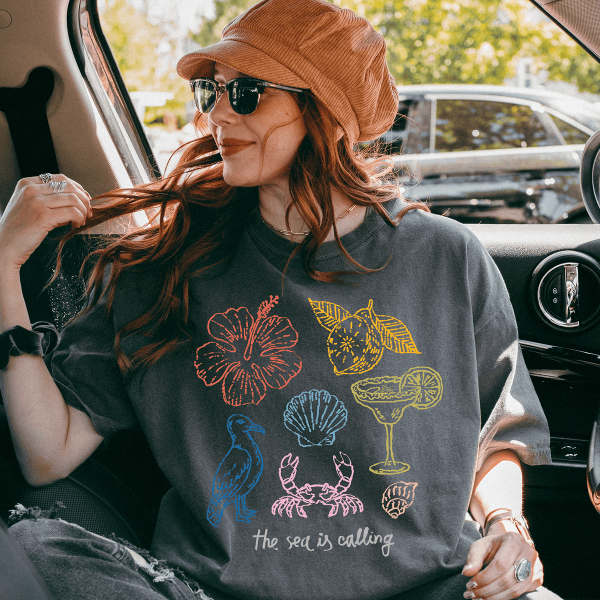 A woman wearing a hat and sunglasses is sitting in a car, looking out the window. She is wearing a gray t-shirt with colorful illustrations of various objects, including a hibiscus flower, a cocktail glass, a crab, and a seashell. The text on the t-shirt says "the sea is calling".