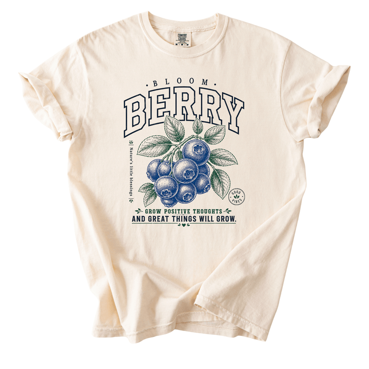 A white t-shirt with a graphic design featuring blueberries and leaves, along with the text "Bloom Berry" and a motivational quote about positive thoughts.