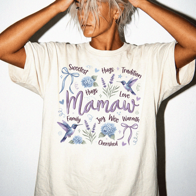 A woman wearing a white t-shirt with a graphic design featuring the word "Mamaw" surrounded by various floral and bird-related elements.