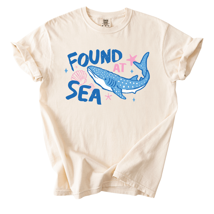 a white t-shirt with a graphic design depicting a blue whale and seashells, along with the text "FOUND AT SEA".