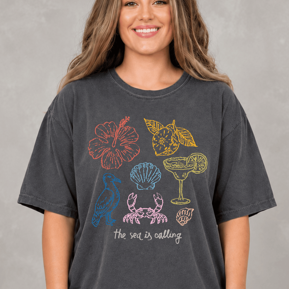 A woman wearing a gray t-shirt with a graphic design featuring various sea-themed illustrations, including a hibiscus flower, a crab, a cocktail glass, and a lemon slice.