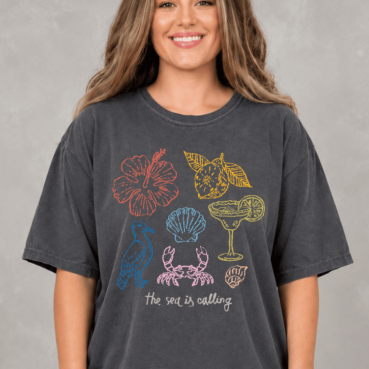 A woman wearing a gray t-shirt with a graphic design featuring various sea-themed illustrations, including a hibiscus flower, a crab, a cocktail glass, and a lemon slice.