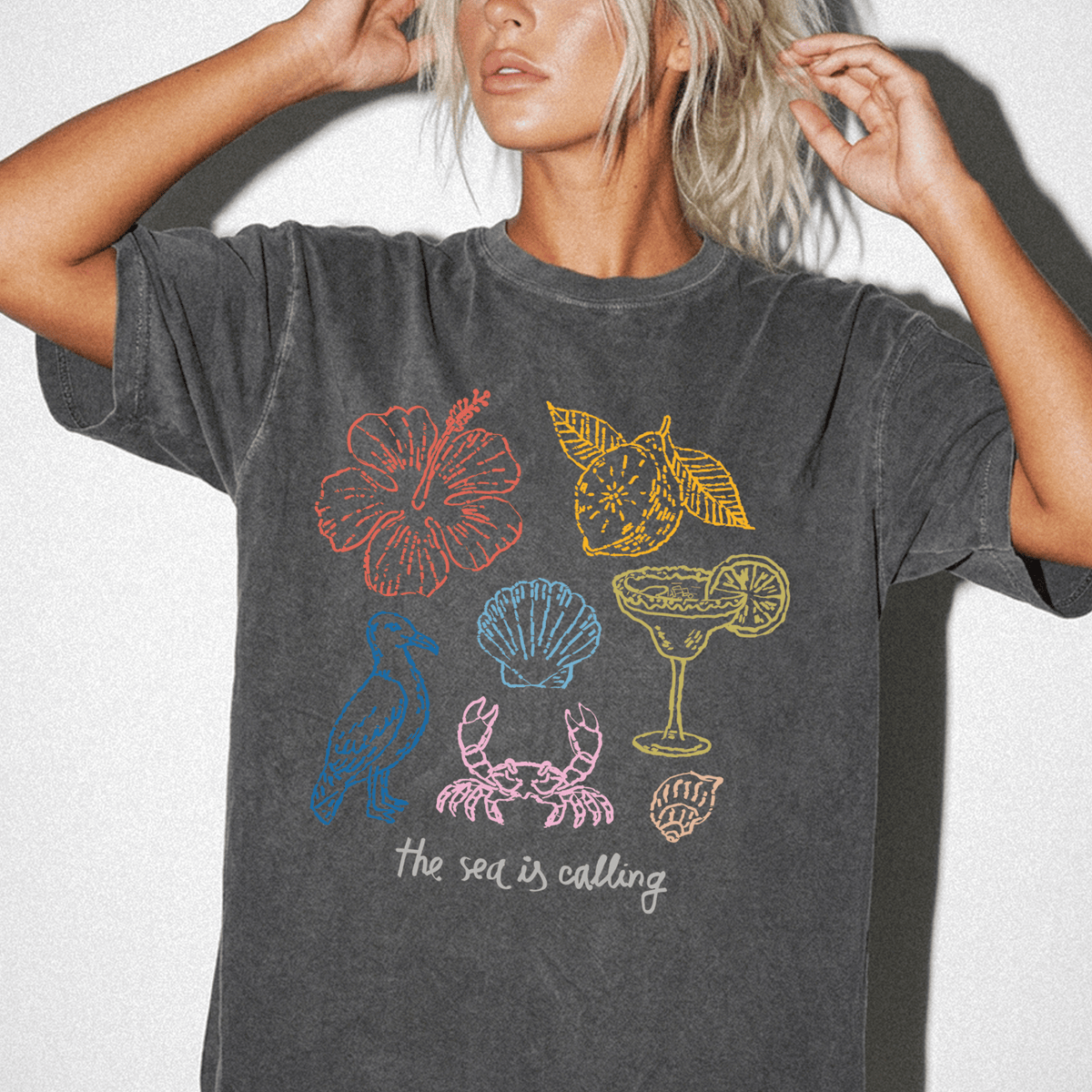 A woman wearing a gray t-shirt with various colorful illustrations of sea-themed objects, including a hibiscus flower, a cocktail glass, and a seashell.