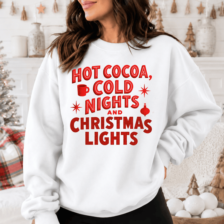 A woman wearing a white sweatshirt with red text that says "HOT COCOA, COLD NIGHTS AND CHRISTMAS LIGHTS".