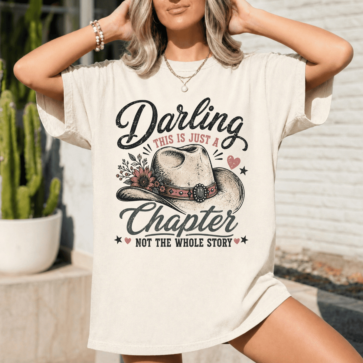 A woman wearing a white t-shirt with a graphic design featuring a cowboy hat and the text "Darling, this is just a chapter, not the whole story".
