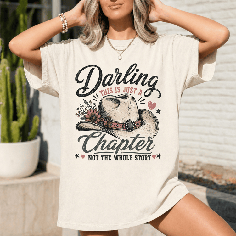 A woman wearing a white t-shirt with a graphic design featuring a cowboy hat and the text "Darling, this is just a chapter, not the whole story".