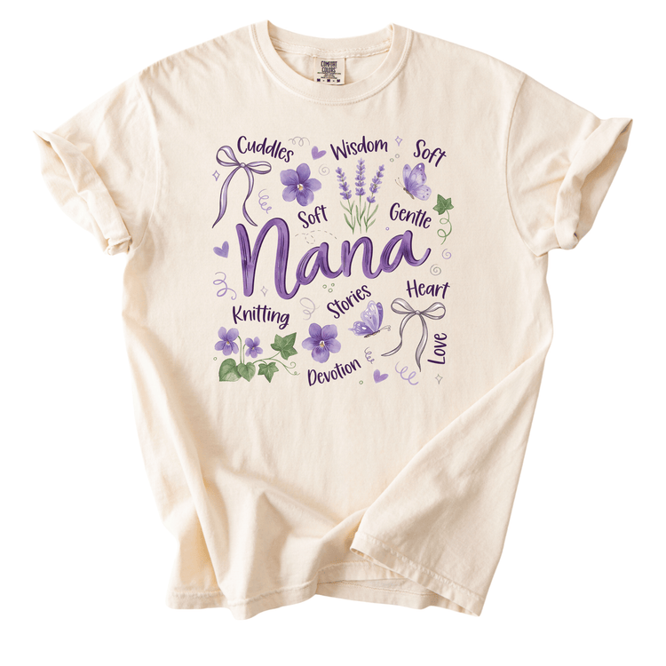 A white t-shirt with a graphic design featuring the word "Nana" surrounded by various purple flowers and leaves, along with the words "cuddles", "soft", "gentle", "knitting", "stories", "heart", "love", and "devotion".