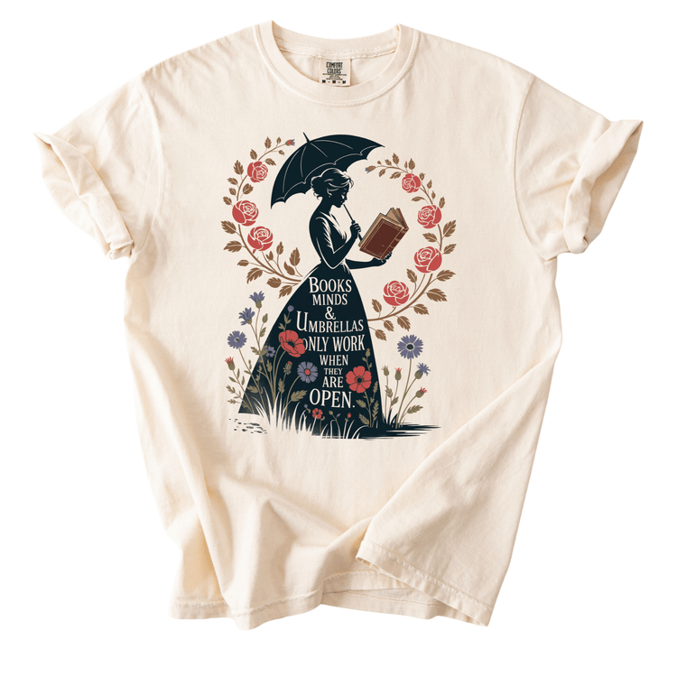 A white t-shirt with a graphic design depicting a woman holding an umbrella and reading a book, surrounded by flowers and a wreath.