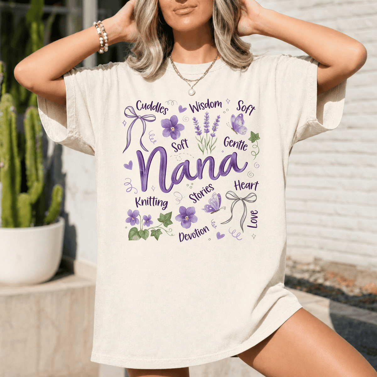 A woman wearing a white t-shirt with a graphic design featuring the word "Nana" surrounded by various purple and green elements, such as flowers, leaves, and ribbons.