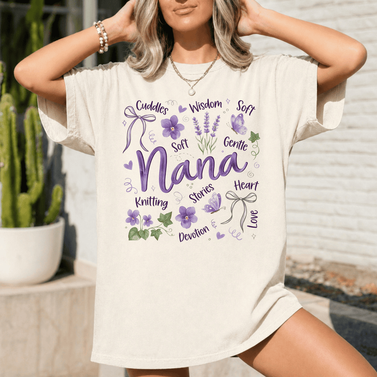 A woman wearing a white t-shirt with a graphic design featuring the word "Nana" surrounded by various purple and green elements, such as flowers, leaves, and ribbons.