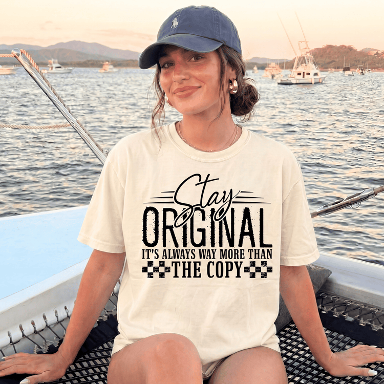 A woman wearing a white t-shirt with a graphic design and text, sitting on a boat in the water with mountains in the background.