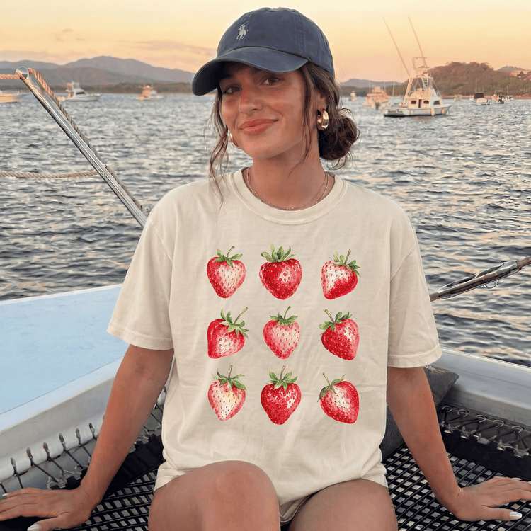 A woman wearing a white t-shirt with a graphic of red strawberries is sitting on a boat, smiling at the camera.