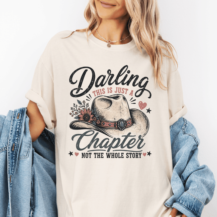 A woman wearing a white t-shirt with a graphic design featuring a cowboy hat and the text "Darling, this is just a Chapter, not the whole story".