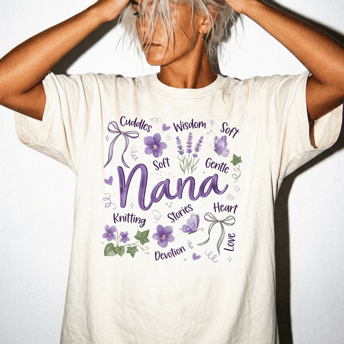 A woman wearing a white t-shirt with a graphic design featuring the word "Nana" surrounded by various purple and green elements, such as flowers, ribbons, and leaves.