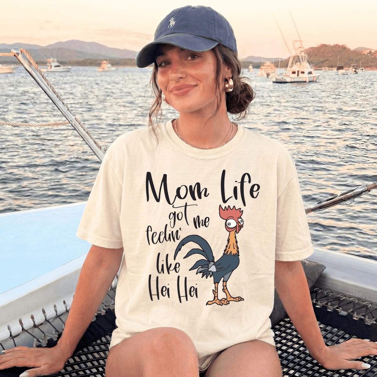 A woman wearing a white t-shirt with a graphic of a rooster and the text "Mom life got me feeling like Hei Hei" is sitting on a boat in the water.