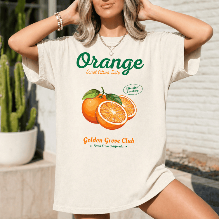A woman wearing a white t-shirt with an orange graphic and text, standing in front of a brick wall.