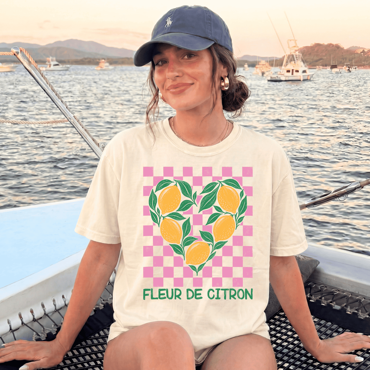 A woman wearing a cap and a white t-shirt with a heart-shaped graphic of lemons is sitting on a boat, smiling at the camera.