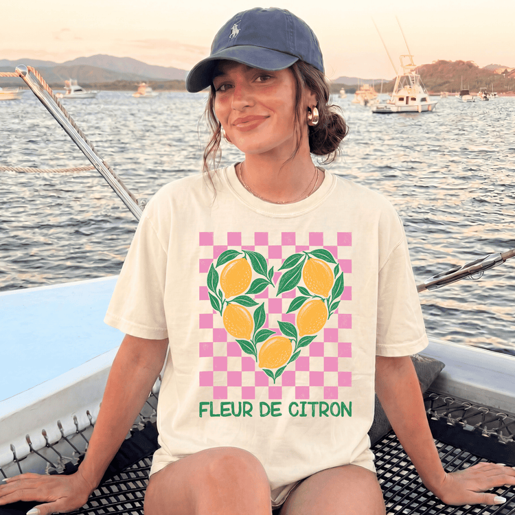 A woman wearing a cap and a white t-shirt with a heart-shaped graphic of lemons is sitting on a boat, smiling at the camera.
