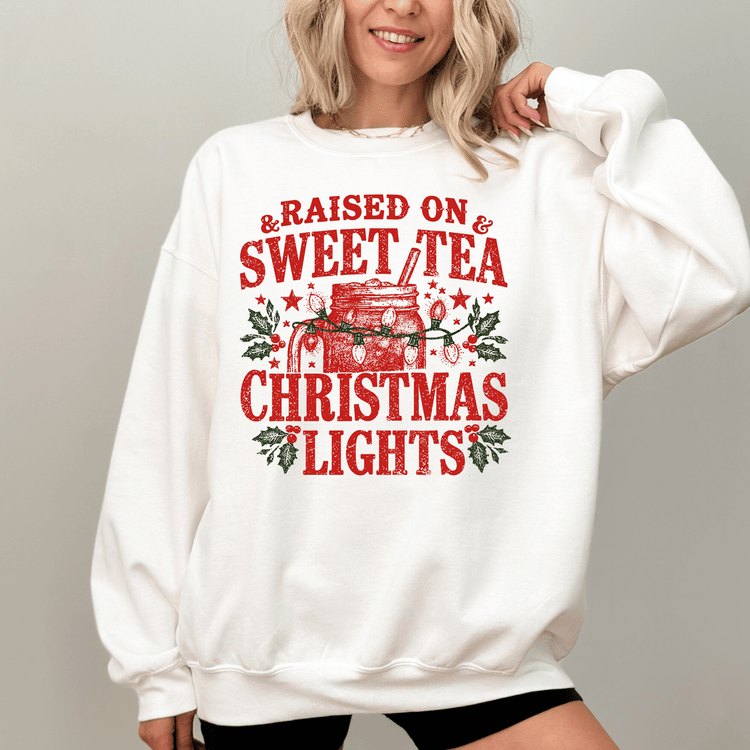 A woman wearing a white sweatshirt with a red and green graphic design that says "Raised on & Sweet Tea Christmas Lights".