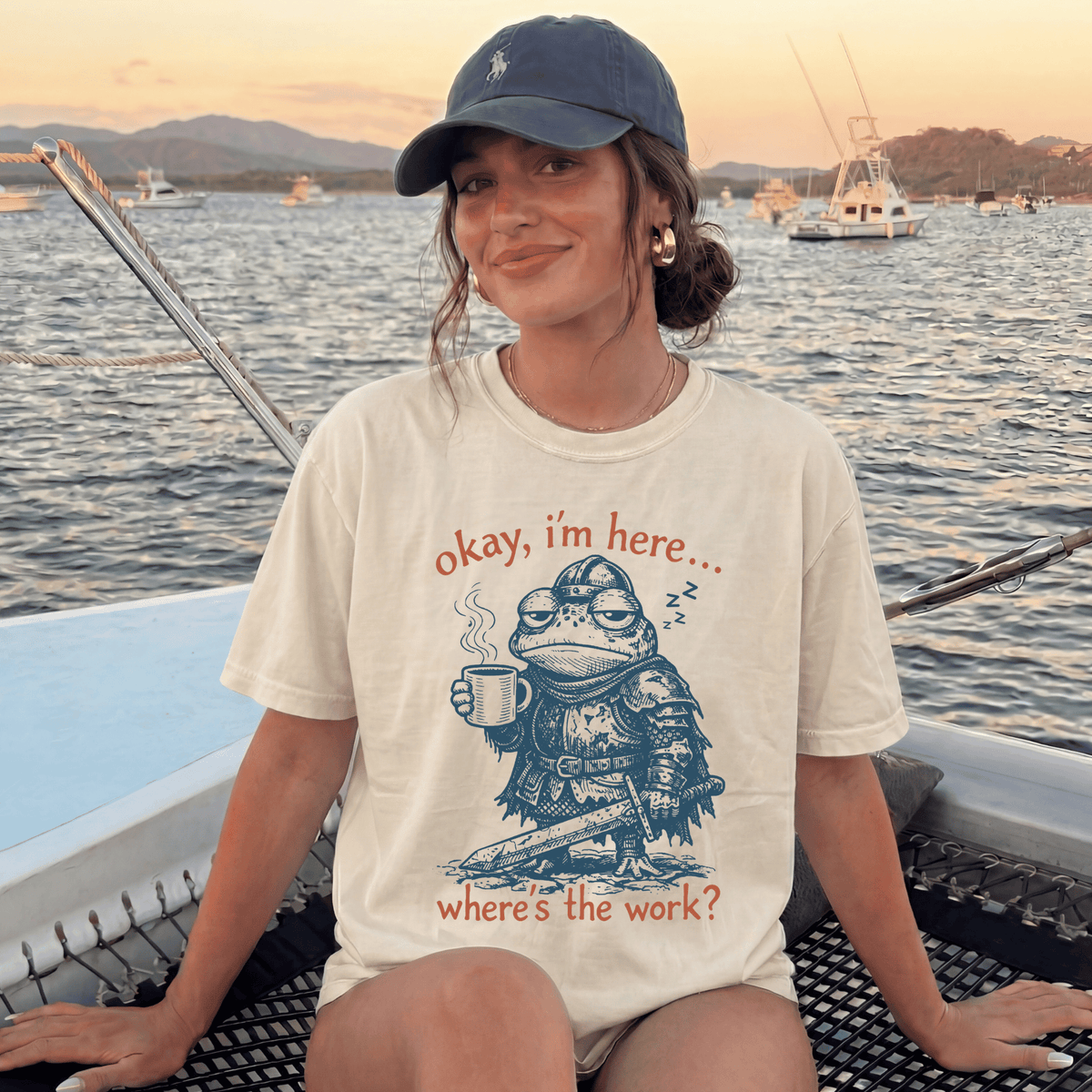 A woman wearing a baseball cap and a t-shirt with a frog character is sitting on a boat, with a sunset in the background.