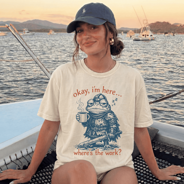A woman wearing a baseball cap and a t-shirt with a frog character is sitting on a boat, with a sunset in the background.