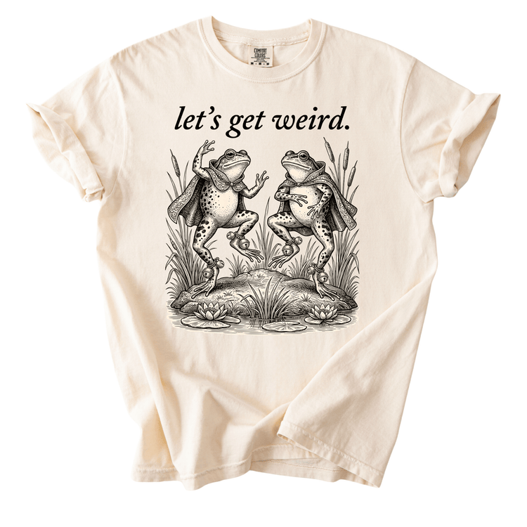 A white t-shirt with a black and white illustration of two frogs standing on rocks and surrounded by plants. Above the illustration, the text "let's get weird." is displayed.