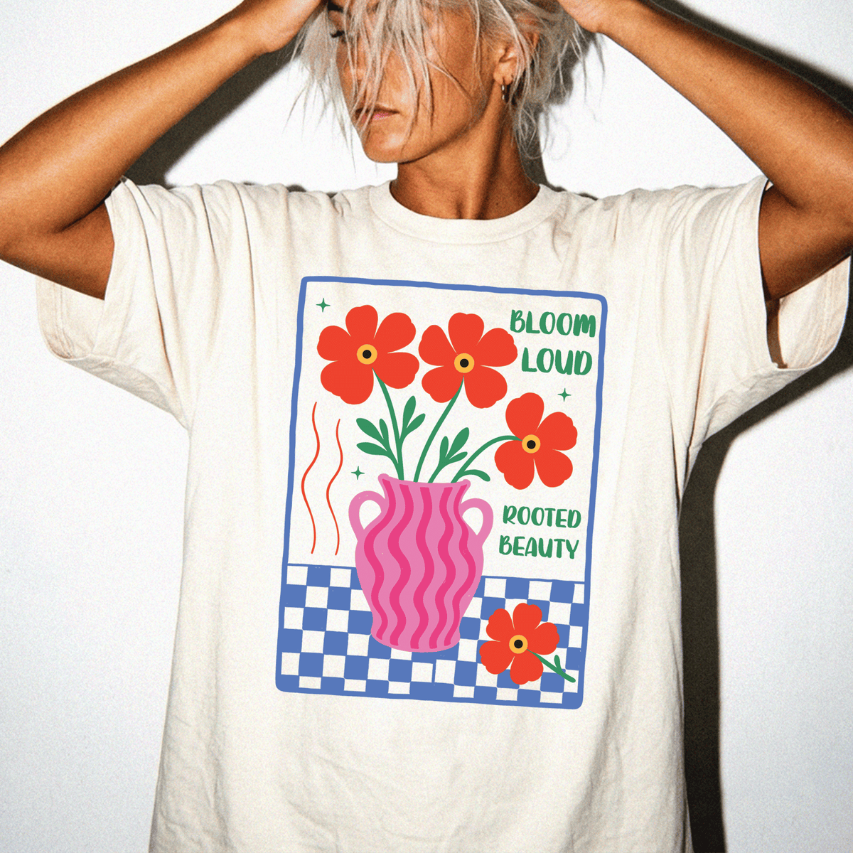 A person wearing a white t-shirt with a graphic design featuring a vase with flowers and the text "Bloom loud rooted beauty".