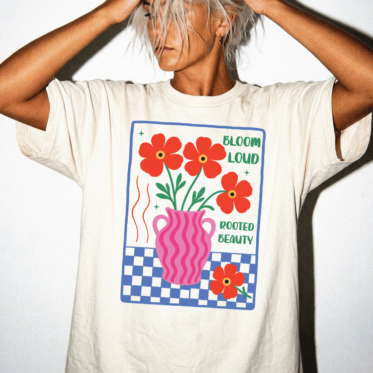 A person wearing a white t-shirt with a graphic design featuring a vase with flowers and the text "Bloom loud rooted beauty".
