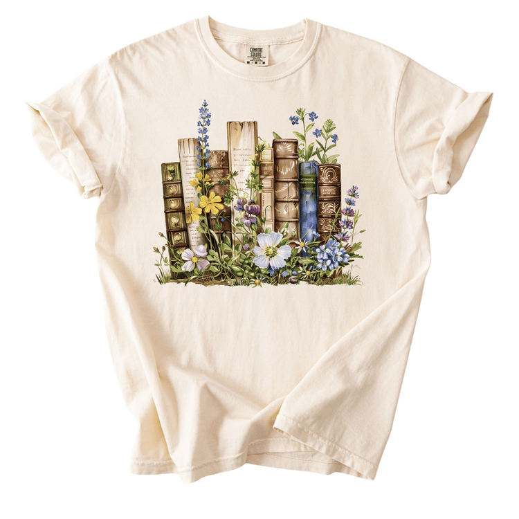 A white t-shirt with a printed design depicting a row of colorful books surrounded by flowers and plants.