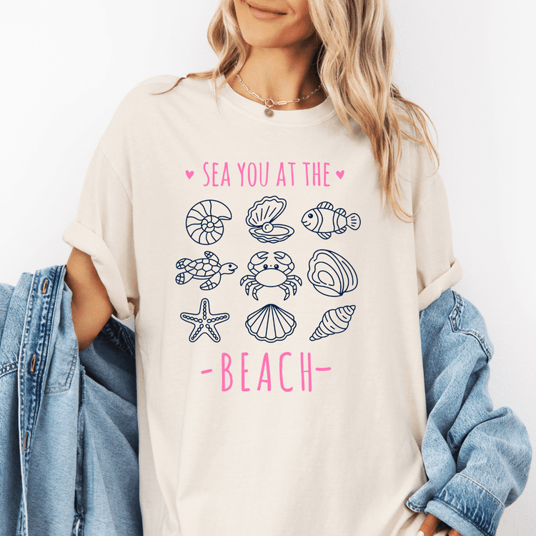 A woman wearing a white t-shirt with a graphic design of various sea creatures and the text "SEA YOU AT THE BEACH" printed on it.
