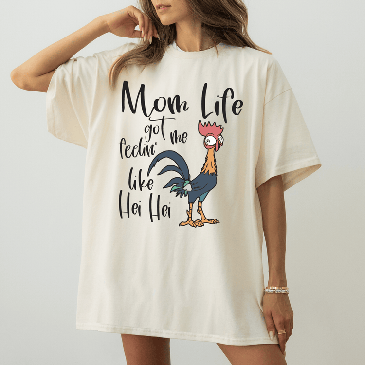 A woman wearing a white t-shirt with a graphic design of a rooster and the text "Mom Life got me feelin' like Her Hei Hei" printed on it.