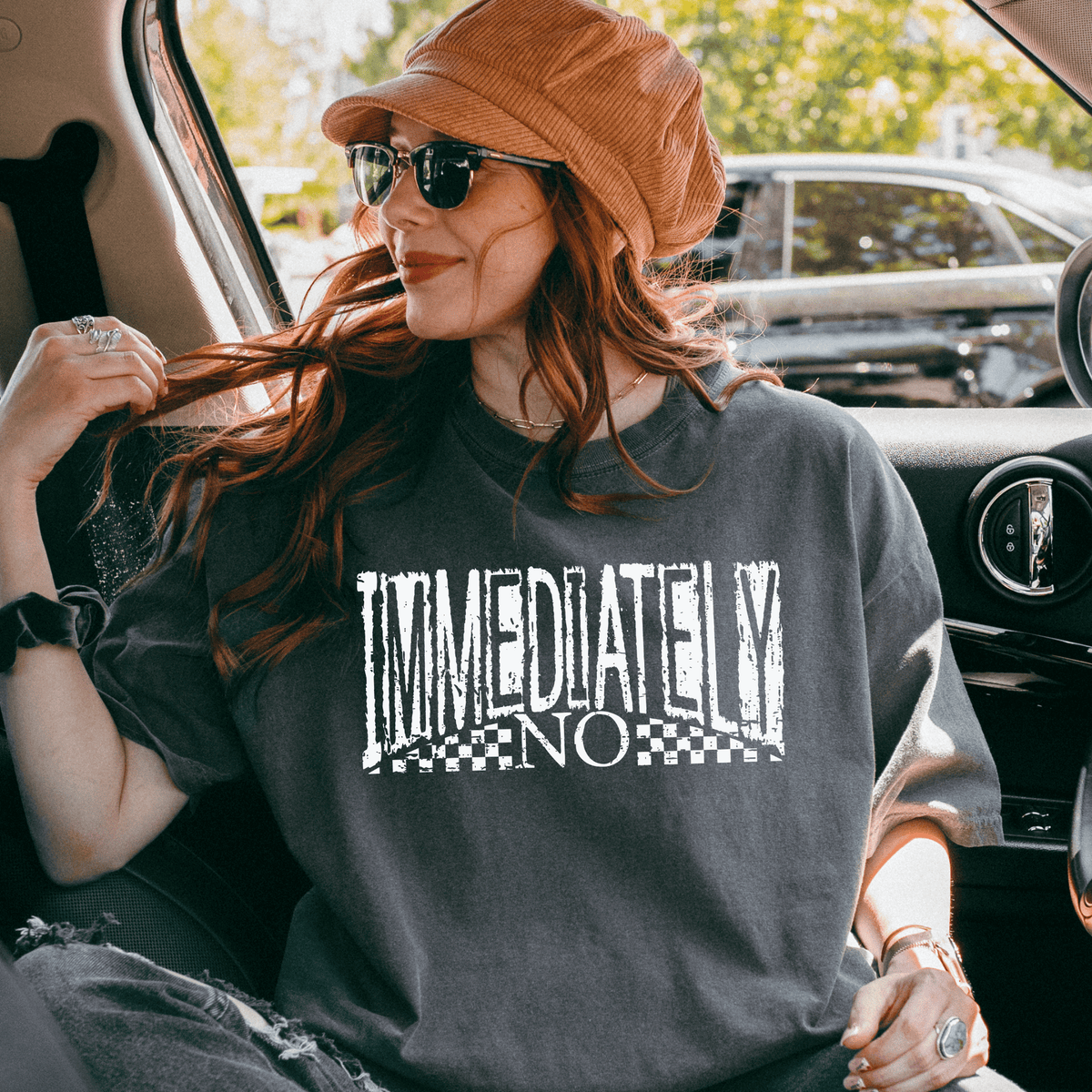 A woman with long red hair wearing a hat and sunglasses is sitting in the back seat of a car, looking out the window. She is wearing a gray t-shirt with the text "IMMEDIATELY NO" printed on it.