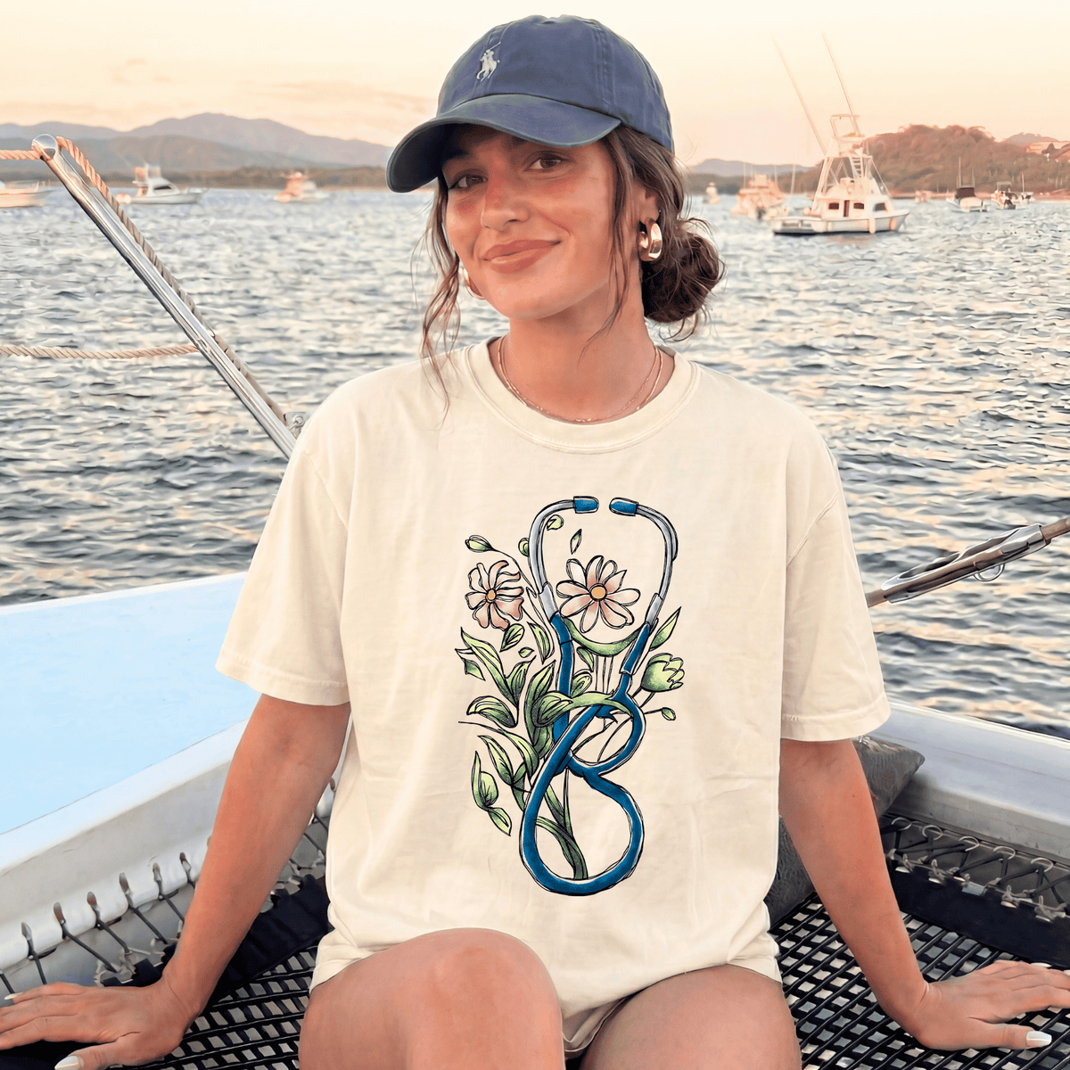 A woman wearing a white t-shirt with a stethoscope design and a flower on it, sitting on a boat with a scenic background of mountains and boats.