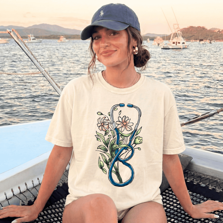 A woman wearing a white t-shirt with a stethoscope design and a flower on it, sitting on a boat with a scenic background of mountains and boats.