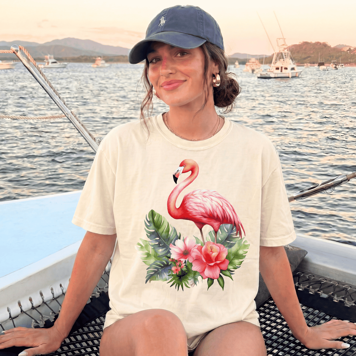 A woman wearing a white t-shirt with a pink flamingo and tropical flowers is sitting on a boat, smiling at the camera.
