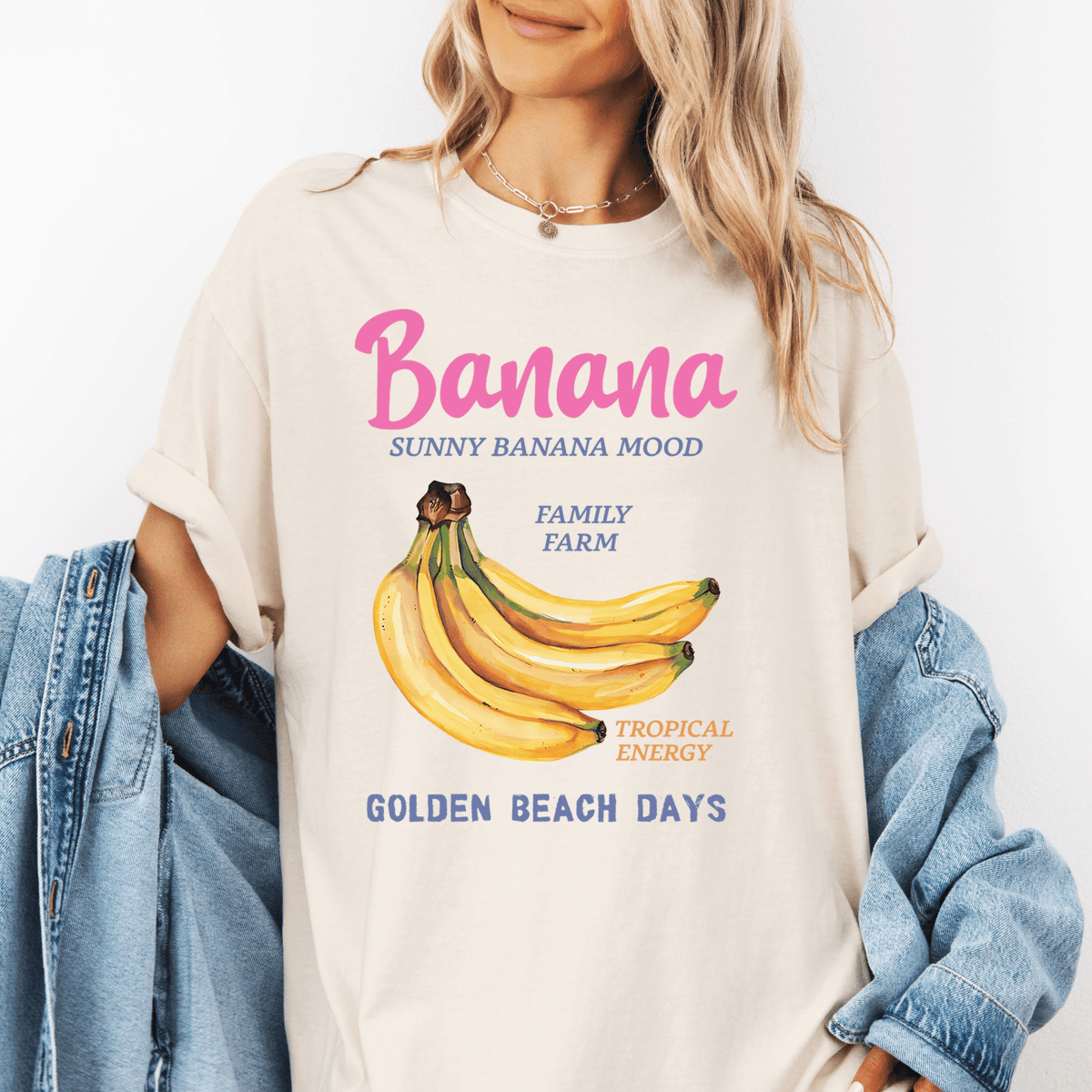 A woman wearing a white t-shirt with a graphic of bananas and text that says "Banana SUNNY BANANA MIND FAMILY FARM TROPICAL ENERGY GOLDEN BEACH DAYS".