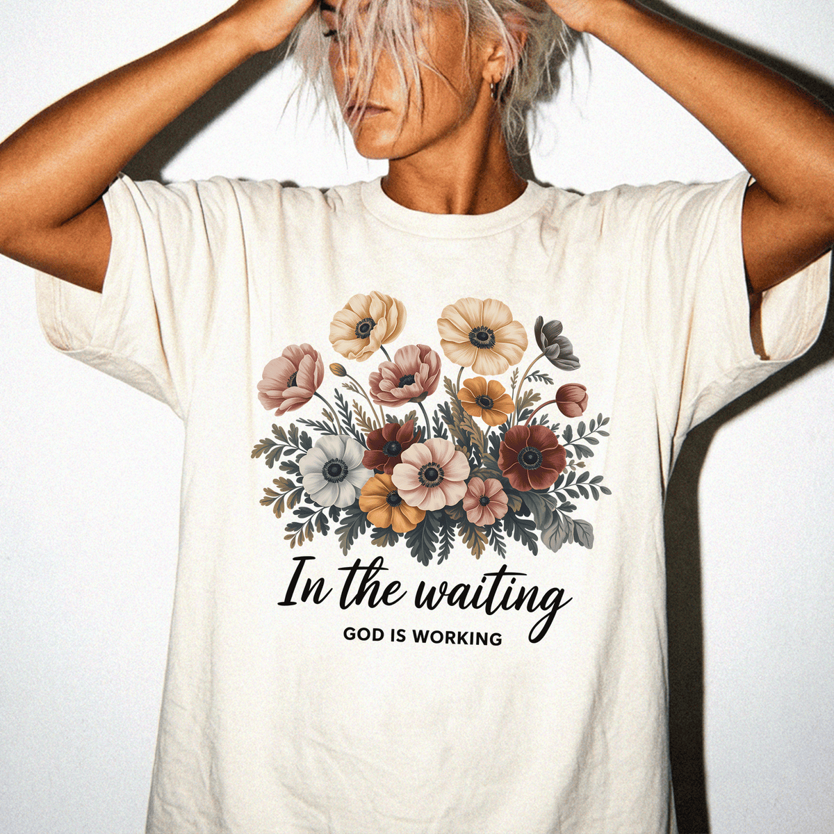 A woman wearing a white t-shirt with a floral design and the text "In the waiting God is working" printed on it.