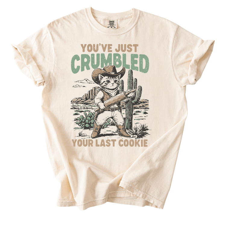 A t-shirt with a graphic design depicting a cat wearing a cowboy hat and holding a cactus, with the text "You've just Crumbled Your Last Cookie" printed on it.