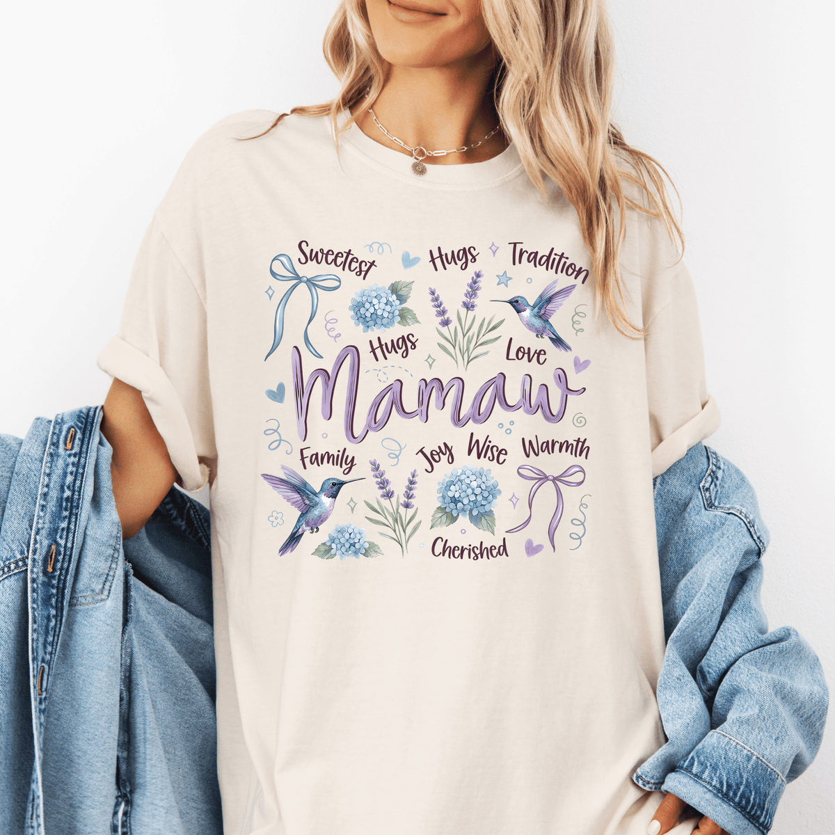 A woman wearing a white t-shirt with a graphic design featuring a bird, flowers, and the word "Mamaw" surrounded by various words such as "Sweetest", "Hugs", "Love", "Family", "Joy Wise", "Warmth", "Charmished", and "Tradition".