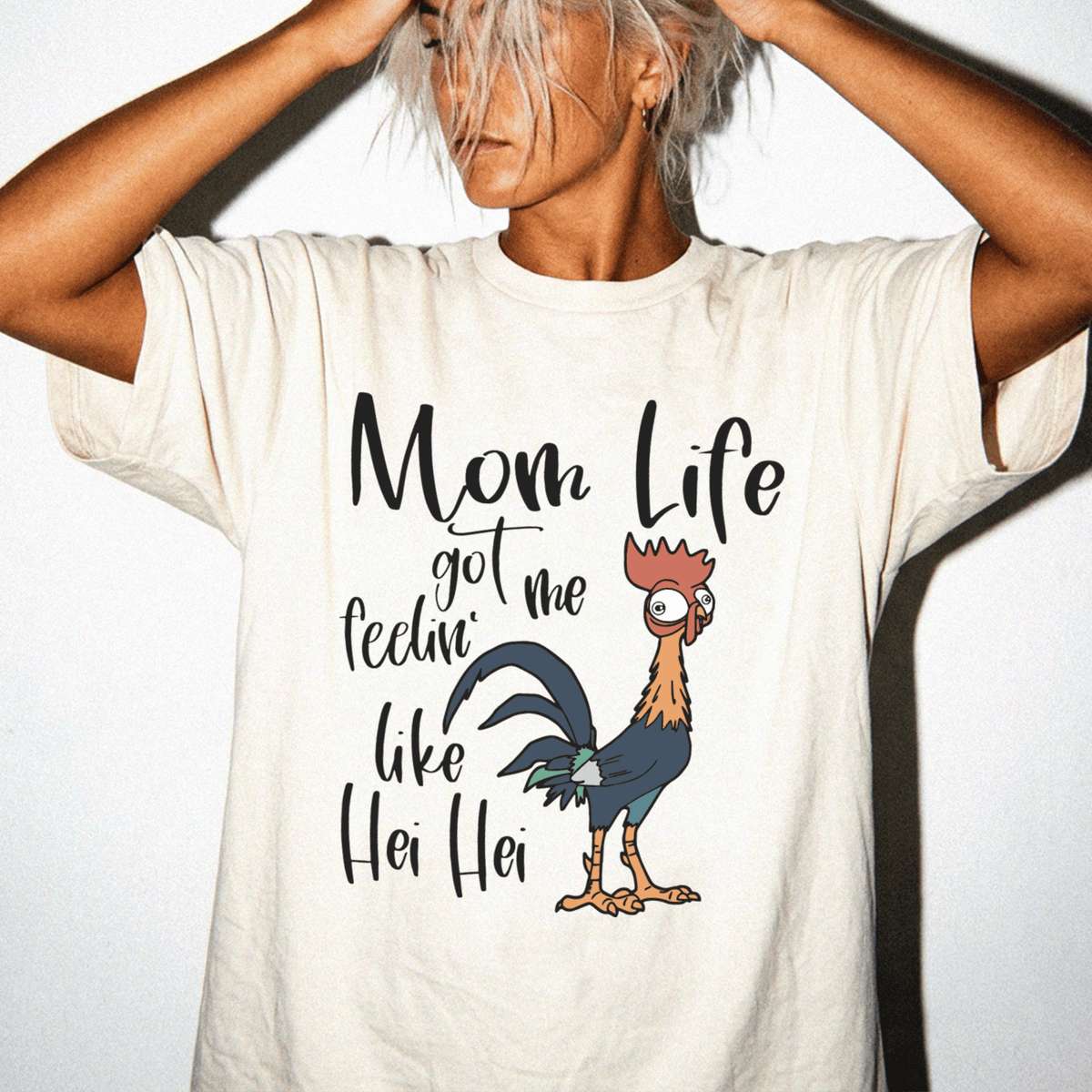 A person wearing a white t-shirt with a graphic design of a rooster and the text "Mom Life got me feelin' like Hei Hei Hei" printed on it.