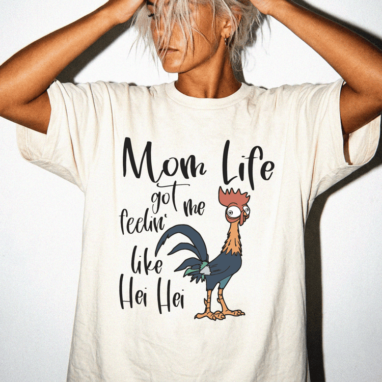 A person wearing a white t-shirt with a graphic design of a rooster and the text "Mom Life got me feelin' like Hei Hei Hei" printed on it.