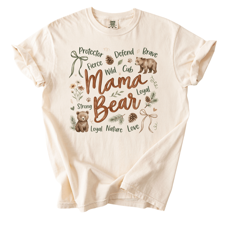 The image is a t-shirt with a design featuring the words "Mama Bear" surrounded by various forest-themed elements such as pine cones, leaves, and a bear.