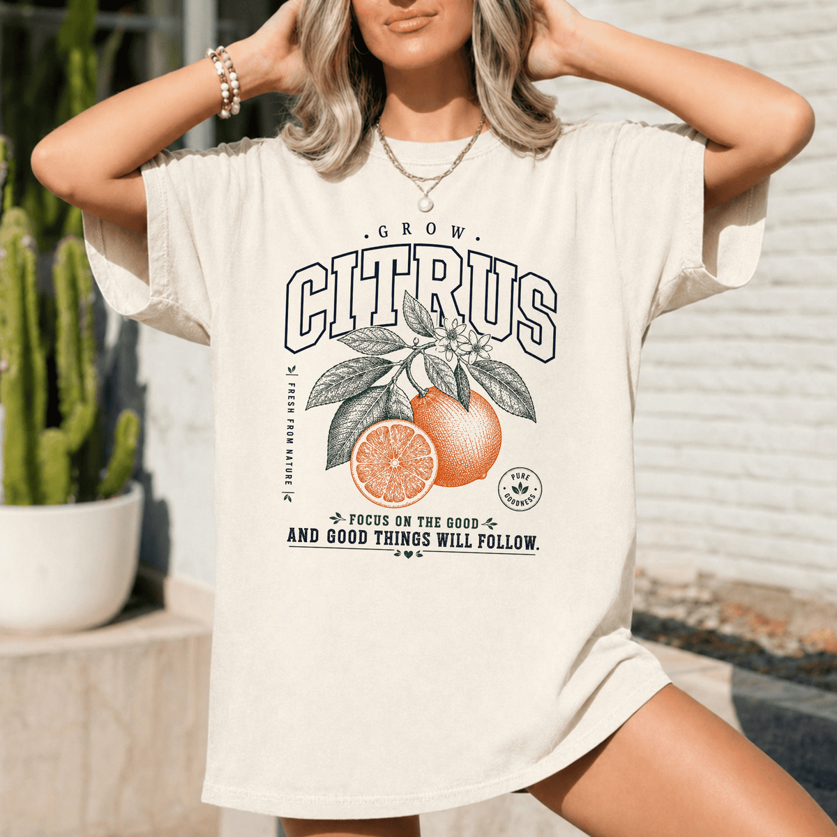 A woman wearing a white t-shirt with a graphic design of an orange and leaves, along with the text "GROW CITRUS" and "FOCUS ON THE GOOD - AND GOOD THINGS WILL FOLLOW".