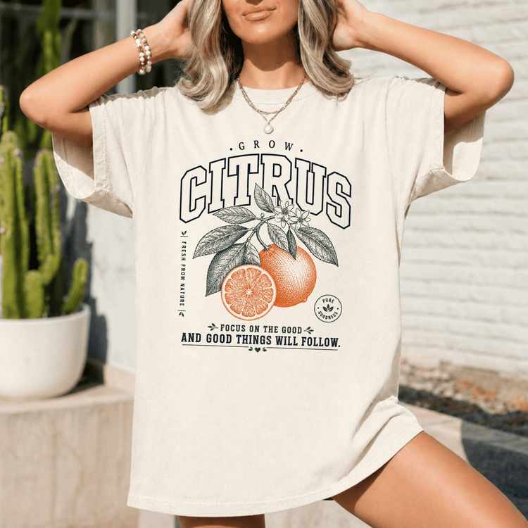 A woman wearing a white t-shirt with a graphic design of an orange and leaves, along with the text "GROW CITRUS" and "FOCUS ON THE GOOD - AND GOOD THINGS WILL FOLLOW".