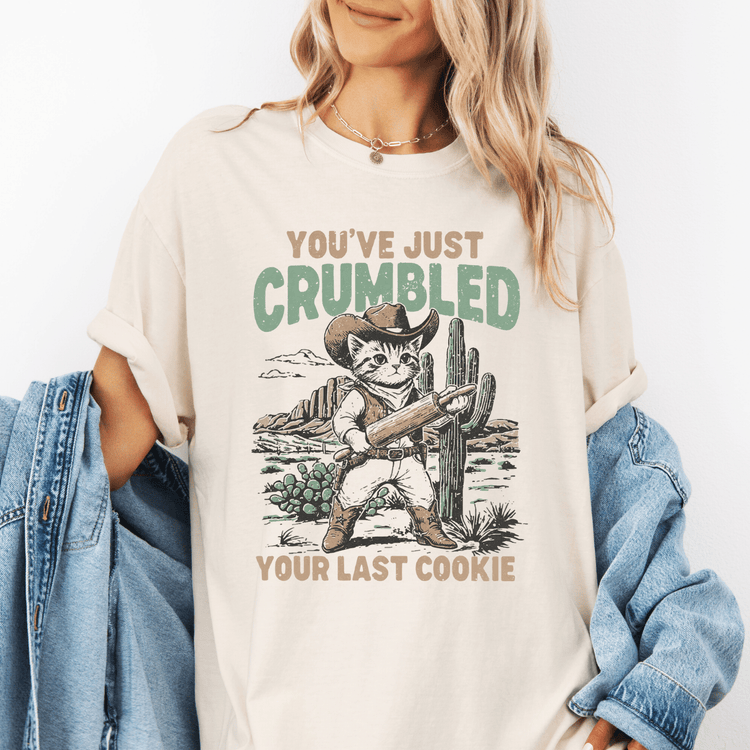 A woman wearing a white t-shirt with a graphic design of a cat holding a cactus and the text "You've just Crumbled Your Last Cookie" printed on it.