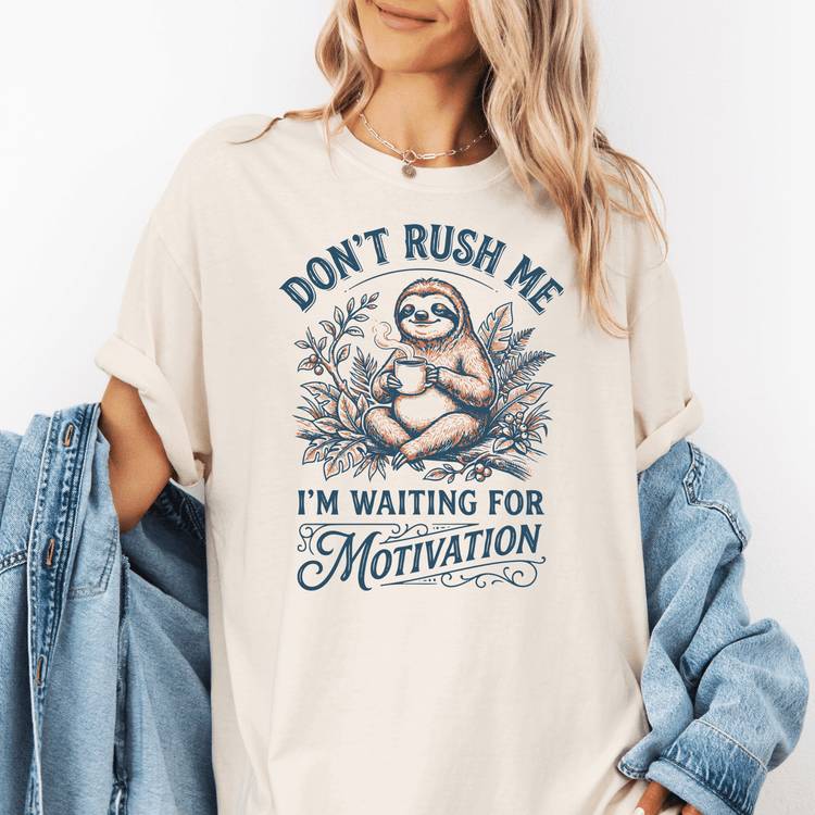 A woman wearing a white t-shirt with a graphic design of a sloth holding a cup of coffee and the text "Don't Rush Me I'm Waiting for Motivation".