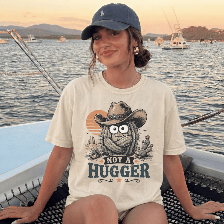 A woman wearing a hat and a t-shirt with a graphic of a cactus wearing a cowboy hat and the text "NOT A HUGGER" is sitting on a boat in the water.