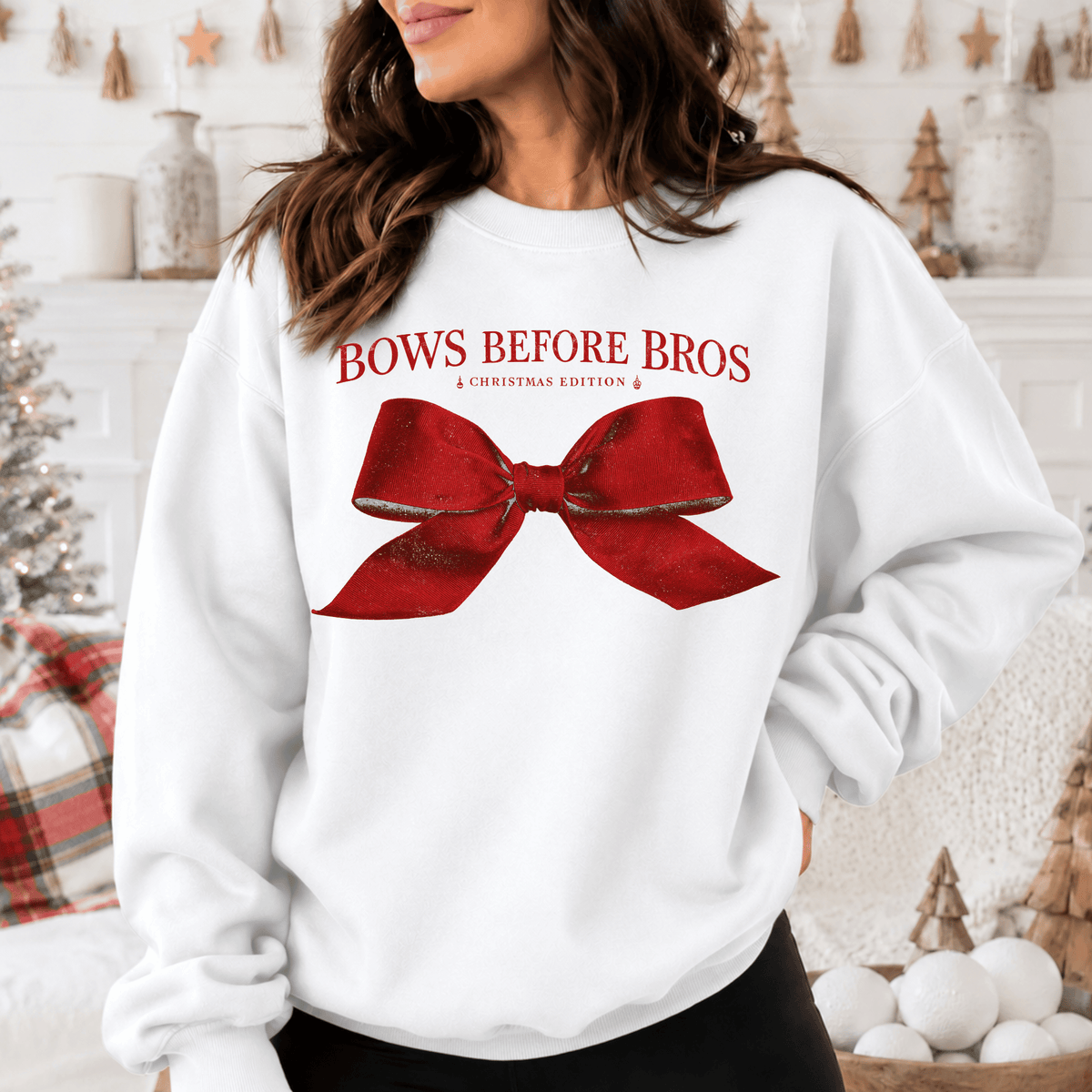 A woman wearing a white sweatshirt with a red bow on it, standing in front of a Christmas tree and other holiday decorations.