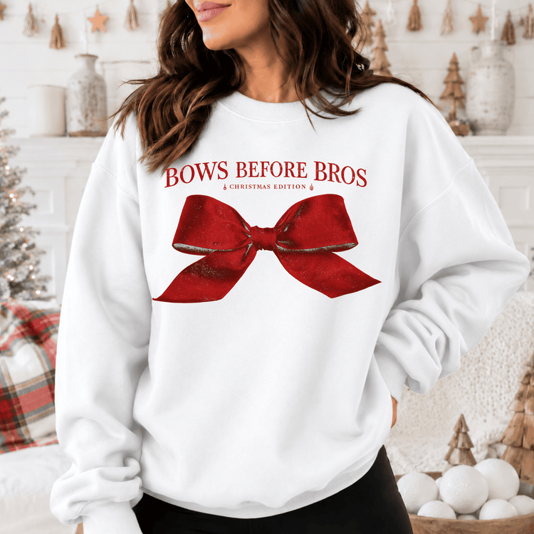 A woman wearing a white sweatshirt with a red bow on it, standing in front of a Christmas tree and other holiday decorations.