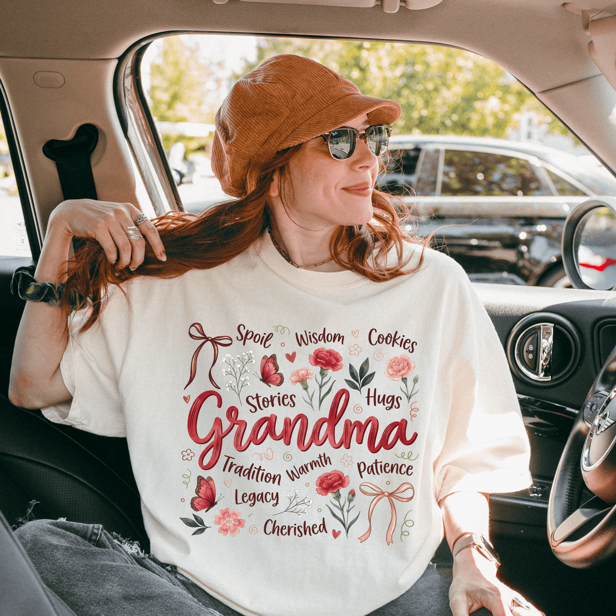 A woman wearing a white t-shirt with the word "Grandma" and various words such as "Spool", "Wise", "Cookies", "Hugs", "Stories", "Hugs", "Legacy", "Patience", "Charm", and "Hugs" printed on it, sitting in a car.